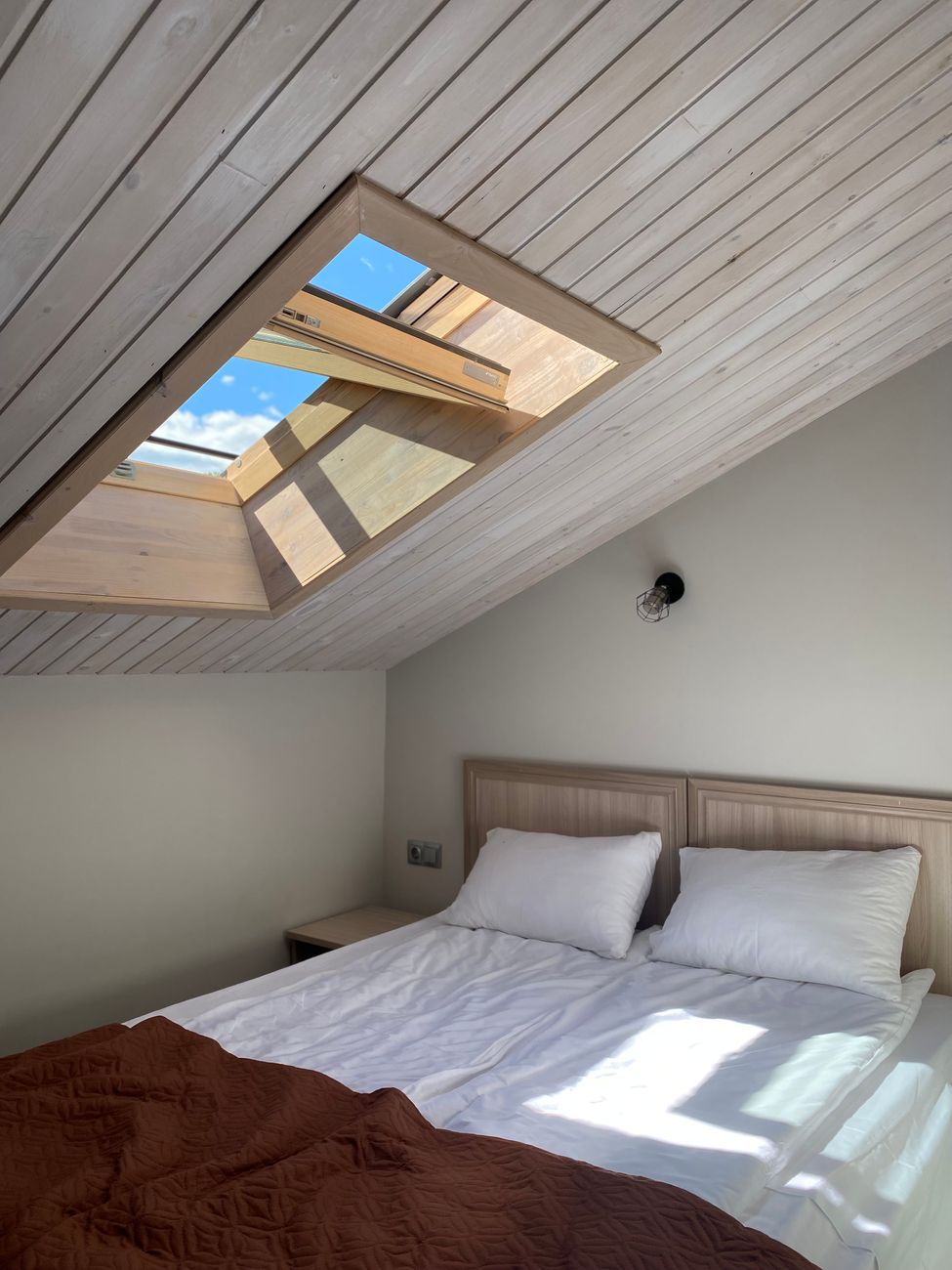 A double bed under a sloped ceiling, positioned beneath a roof window that opens to the sky.