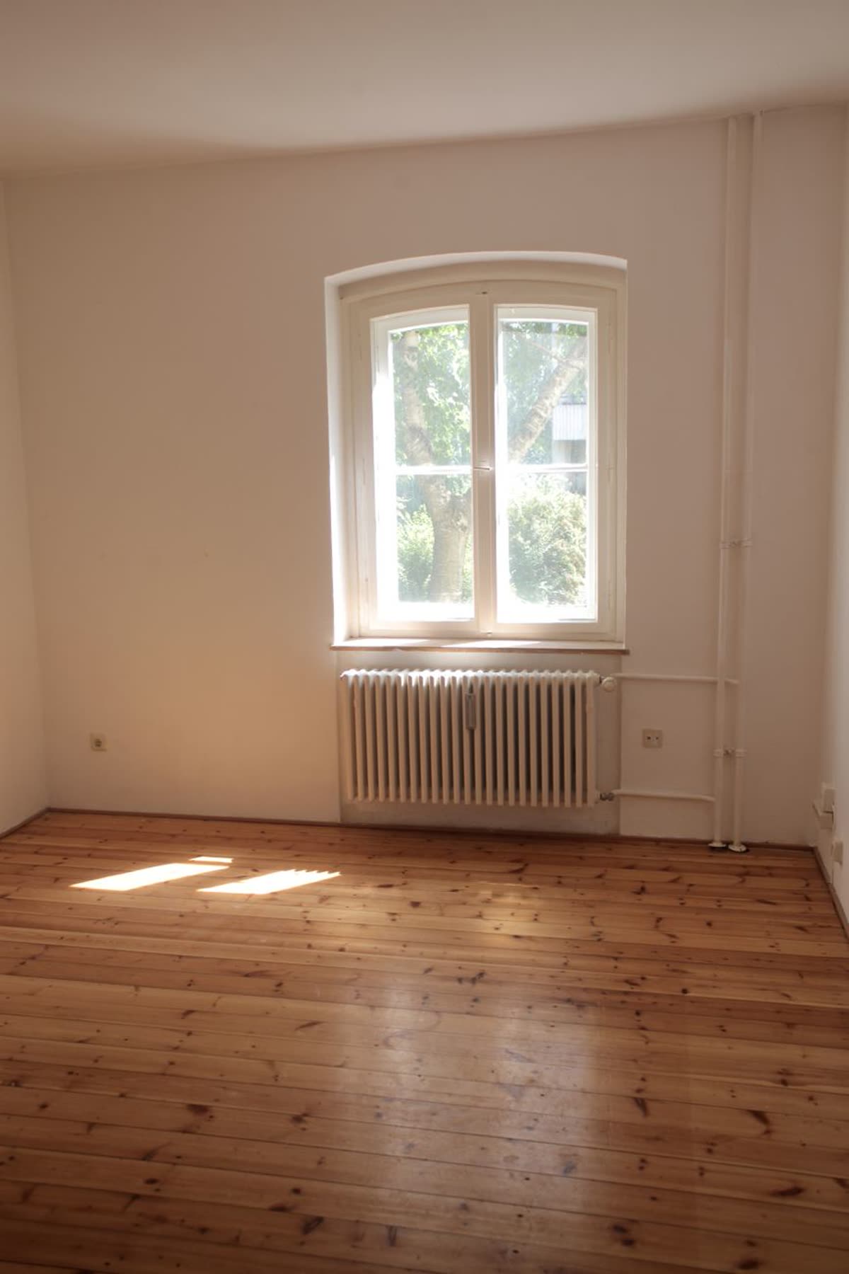 An empty room in an ordinary flat: bare wooden floorboards, a radiator under a single tall window, nothing carried in yet.
