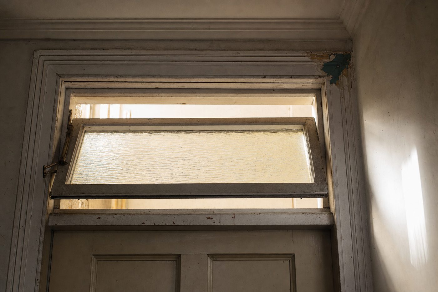 A transom above an interior door, tilted open on its centre pivot, with the brass operator arm visible at one side.